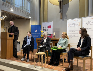 Wilfried Helm am Pult stehend, begrüßt die fünf sitzenden Teilnehmer:innen des Zeitzeugengesprächs im Altarraum der Stadtpfarrkirche. Im Hintergrund der Altar und Rollups der Veranstalter.